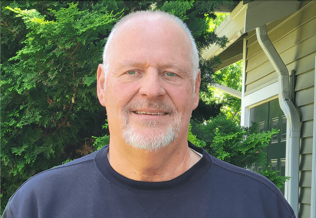 Smiling senior man in navy shirt standing outside home with evergreen shrubs in background
