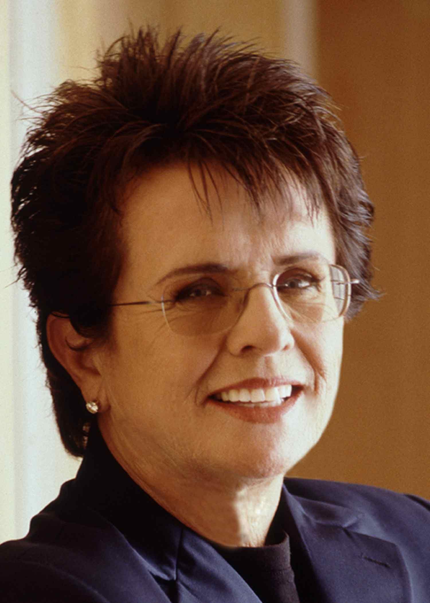 Professional portrait of person with spiky brown hair wearing glasses and navy blazer, smiling warmly at camera