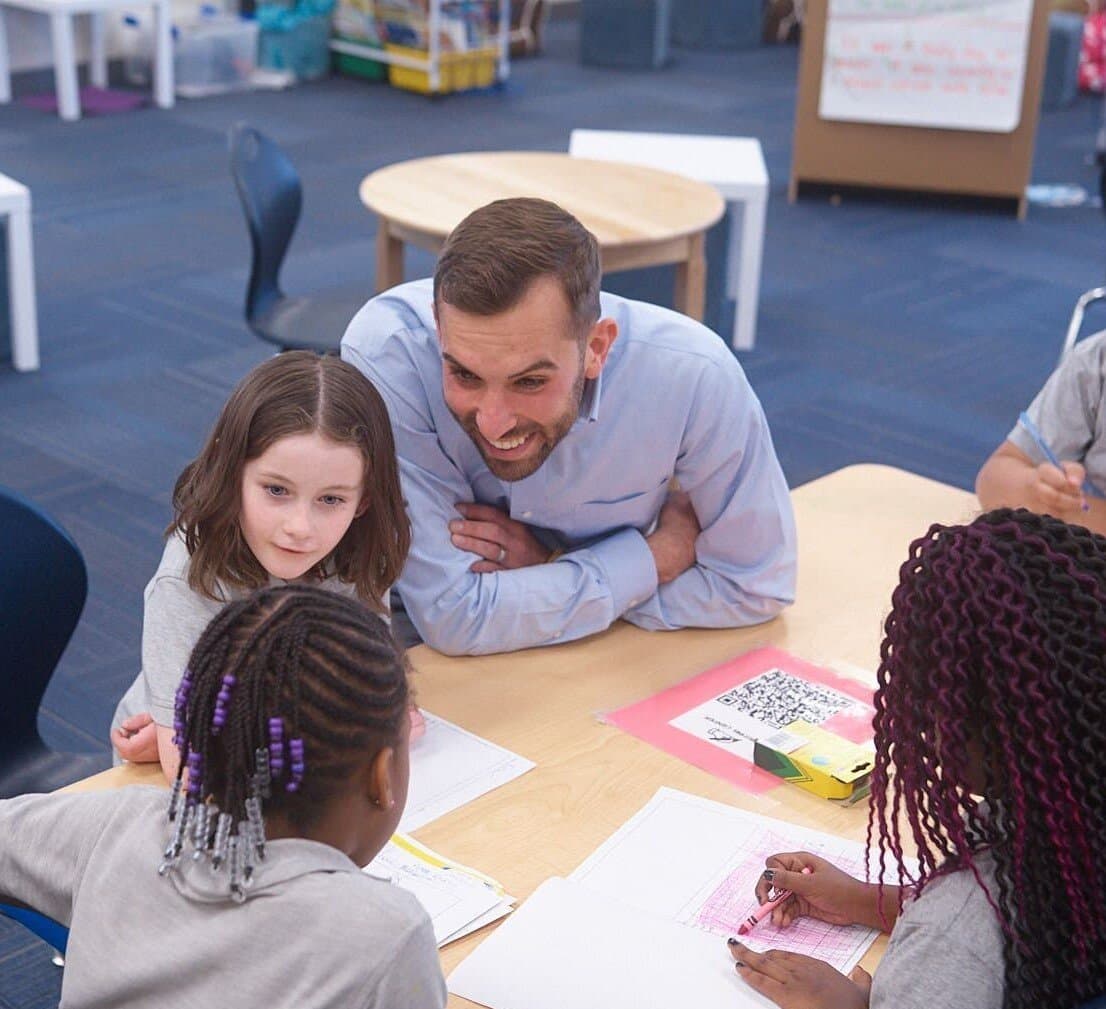 Teacher leaning over table engaging with diverse group of students working on papers in classroom setting