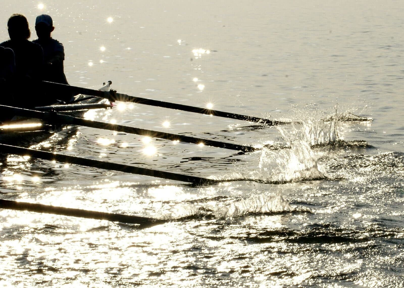 Close-up of rowing oars slicing through sparkling water, creating splashes against bright sunlit reflections