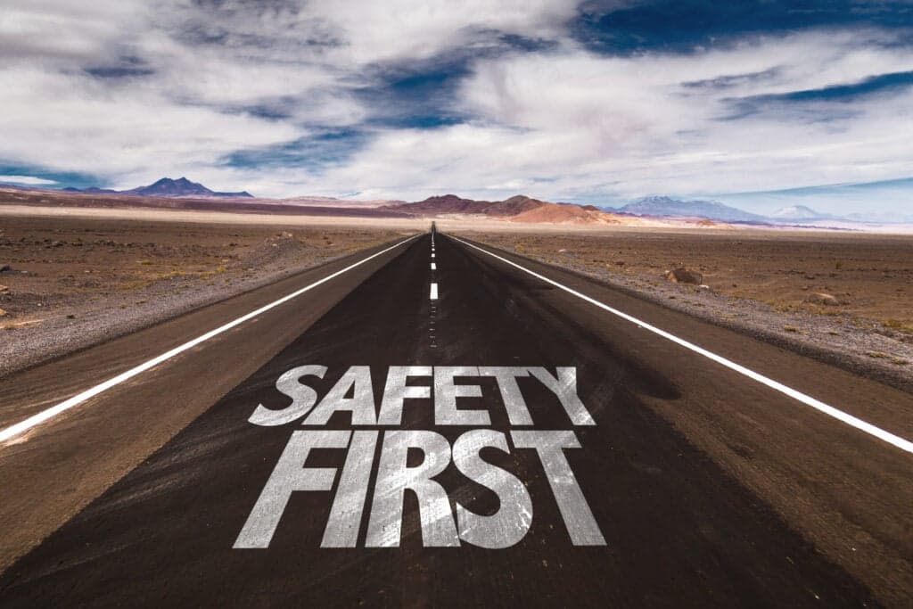 Text 'SAFETY FIRST' painted on an empty desert highway stretching toward mountains under cloudy blue skies