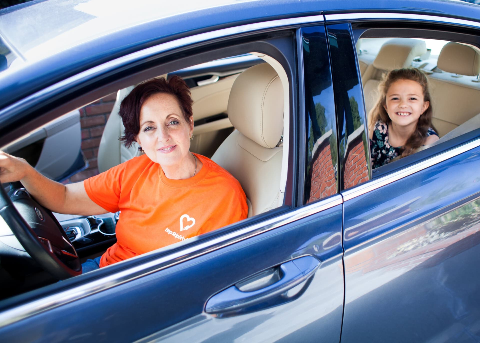 Driver in orange charity t-shirt and young passenger in backseat of blue car, both smiling at camera
