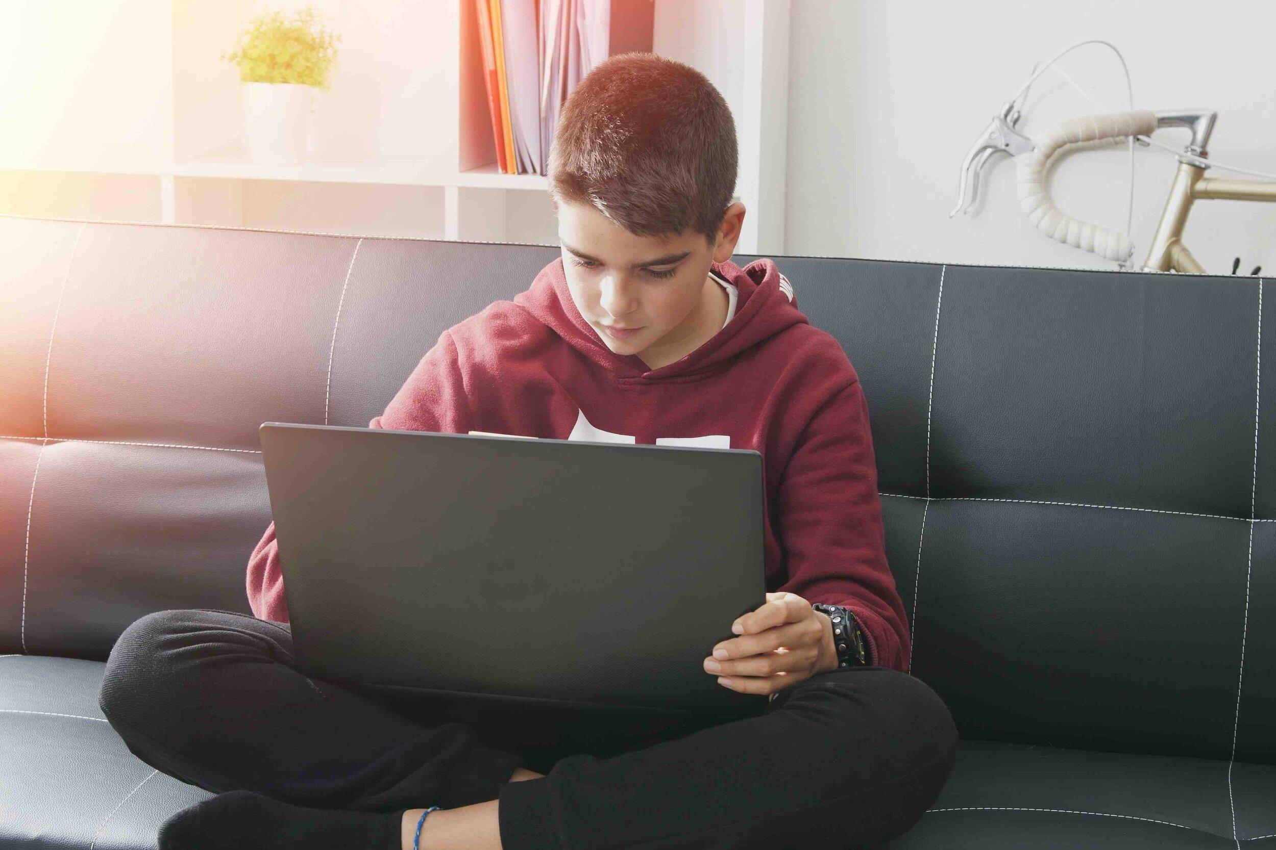 Young teenager in red hoodie sitting cross-legged on couch working on laptop computer