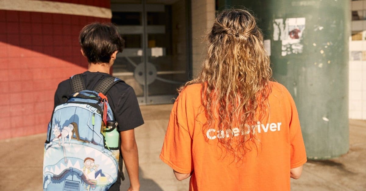 Two students walking, one wearing a Rick and Morty backpack and another in an orange caregiver shirt, viewed from behind