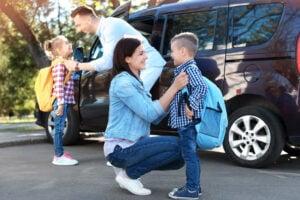Parent kneeling to comfort child at school drop-off while another child stands near family car in the background