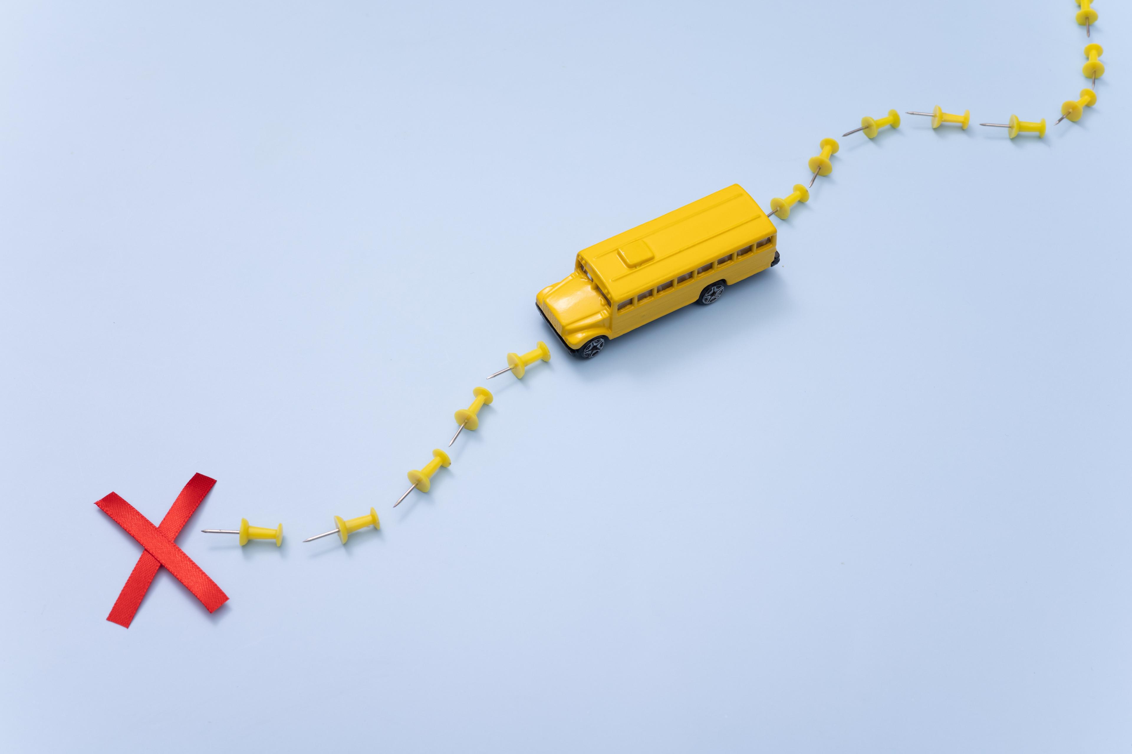 Yellow toy school bus following a path marked by yellow pushpins, leading to a red X mark on light blue background