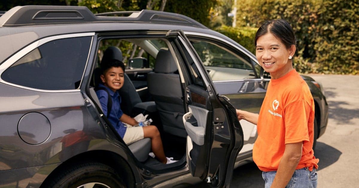 Driver in orange t-shirt standing by car door as young student in school uniform sits in back seat