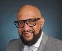 Professional headshot of a man in glasses and grey suit with a friendly smile against a blue backdrop