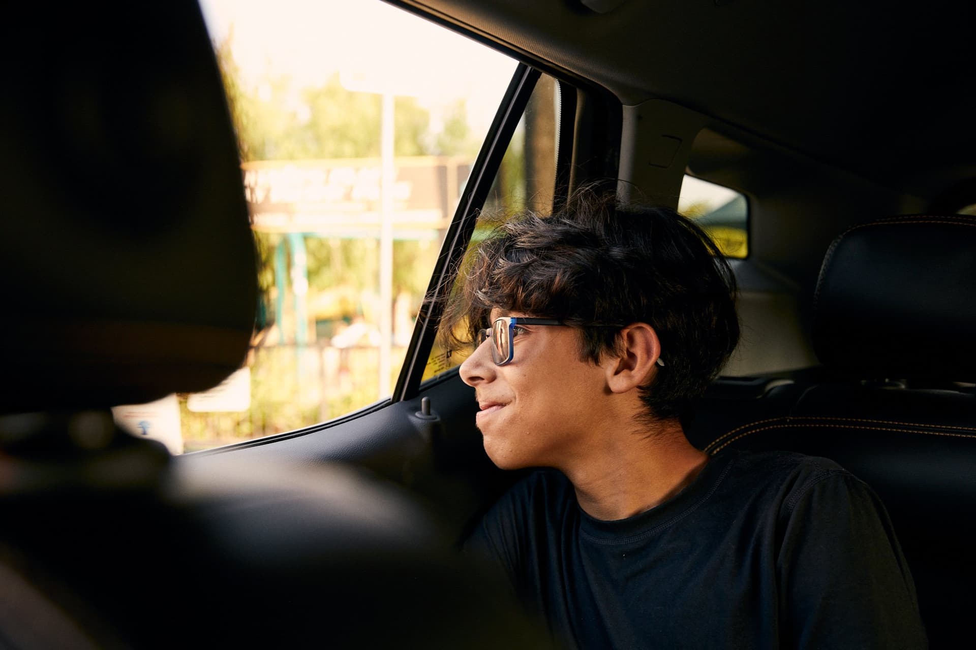 Person wearing glasses in black t-shirt gazing thoughtfully through car window during golden hour