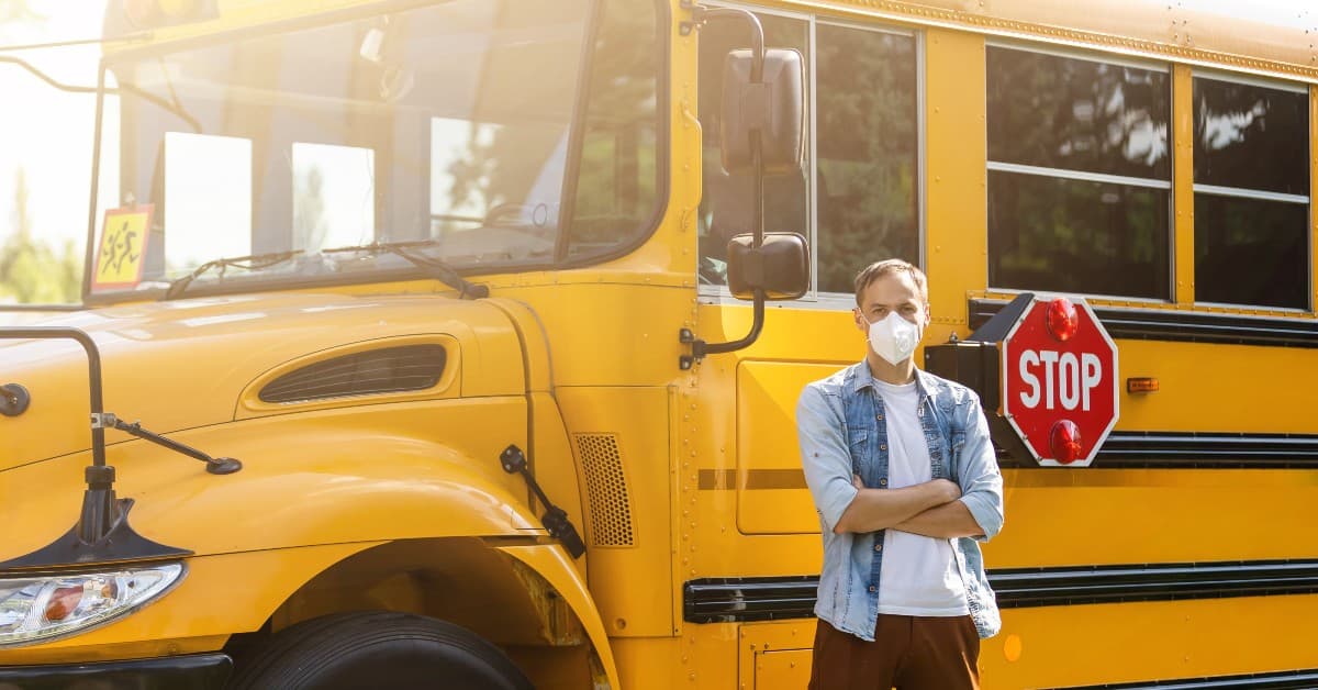 Person wearing face mask and denim jacket standing in front of yellow school bus with stop sign