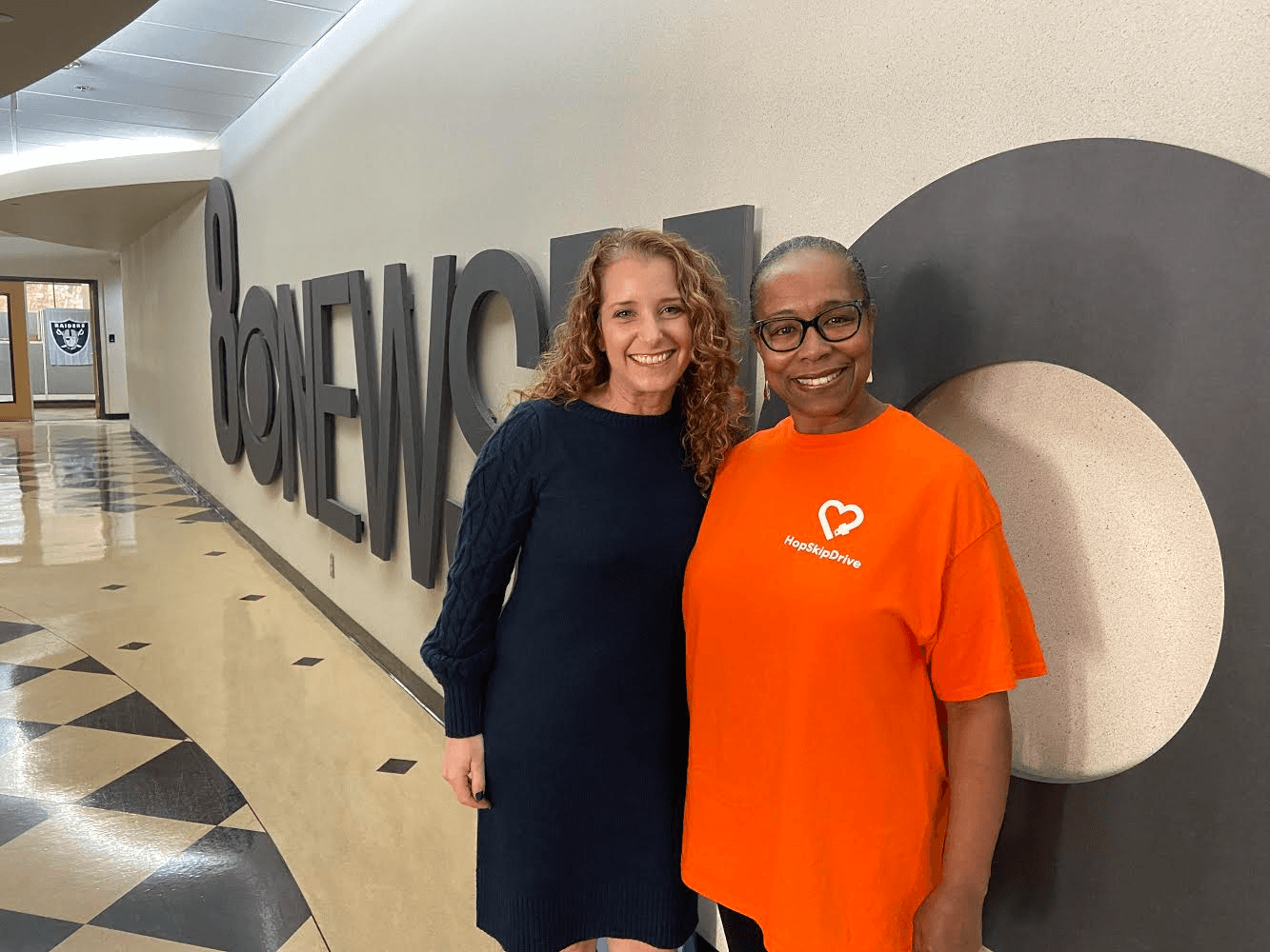 Two professional women standing in front of large wall letters spelling 'BONENO', one in navy dress, one in orange HopSkipDrive t-shirt
