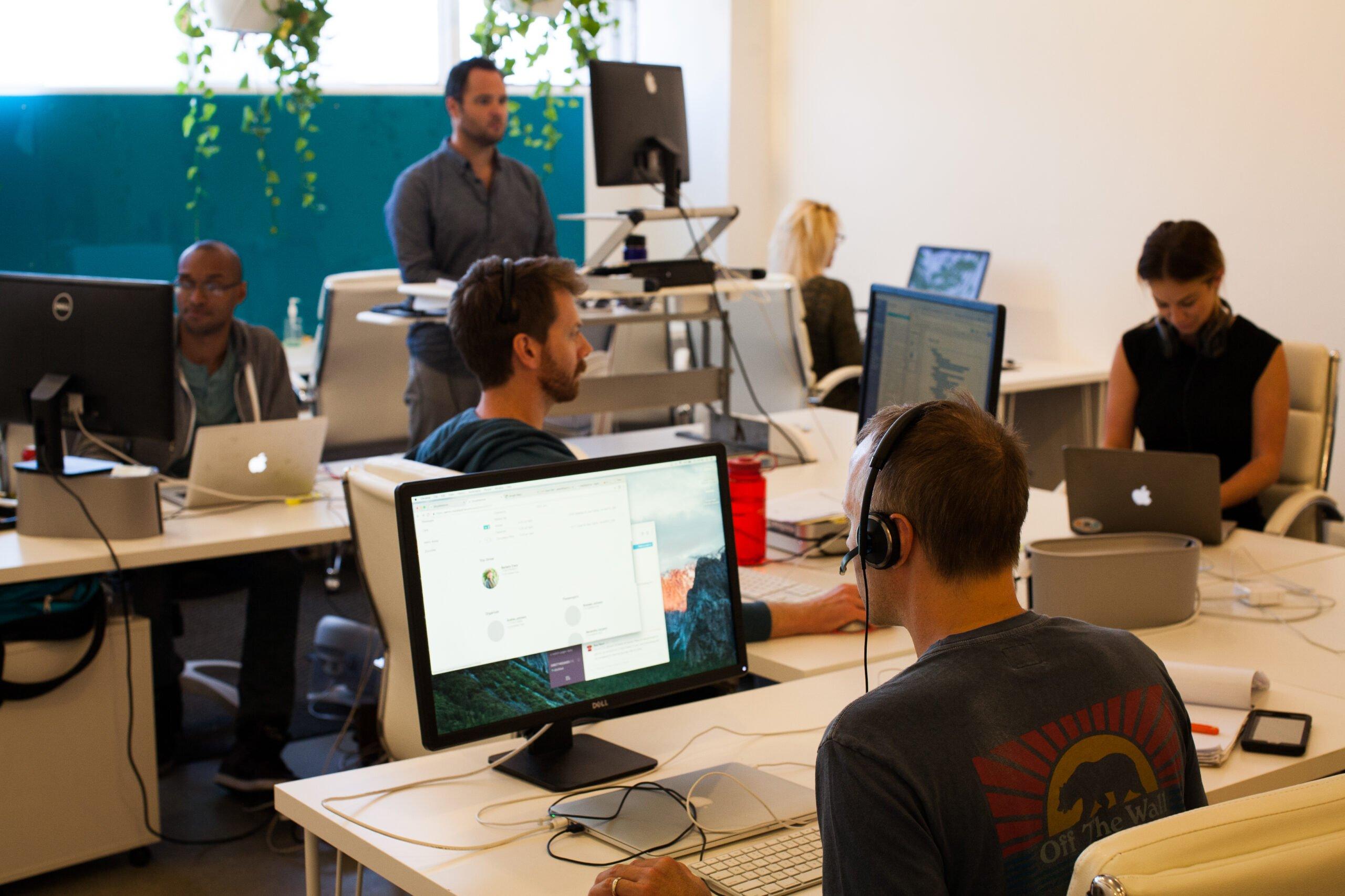 Open office workspace with multiple people working at computers, standing desk setup, and plants decorating the space
