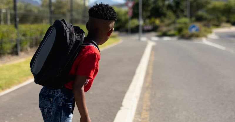 Student in red shirt carrying backpack walking alone on a suburban road with trees and greenery in background