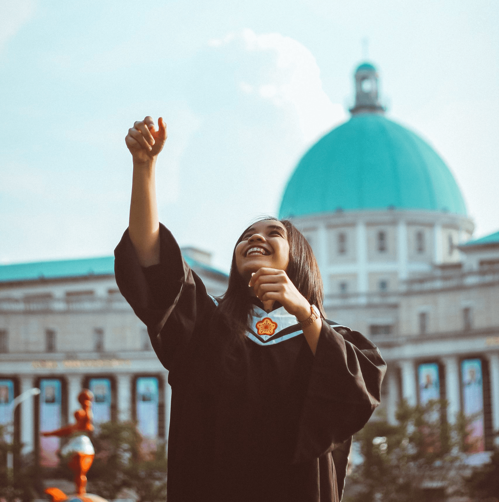 Joyful graduate in black academic gown celebrating with raised arms against backdrop of historic building with turquoise dome