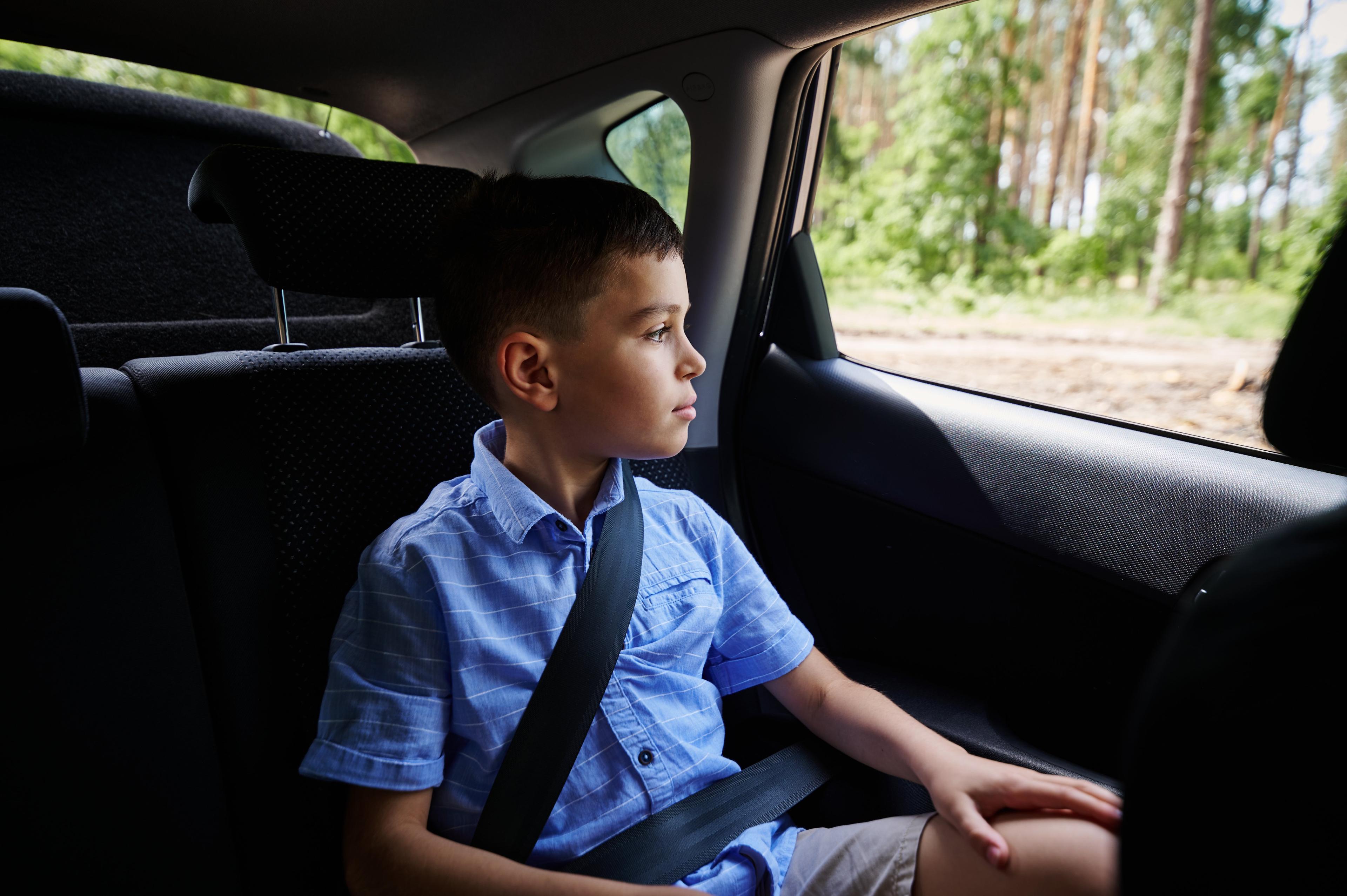 Young boy in blue shirt sitting in car backseat with seatbelt on, gazing thoughtfully out the window at passing trees