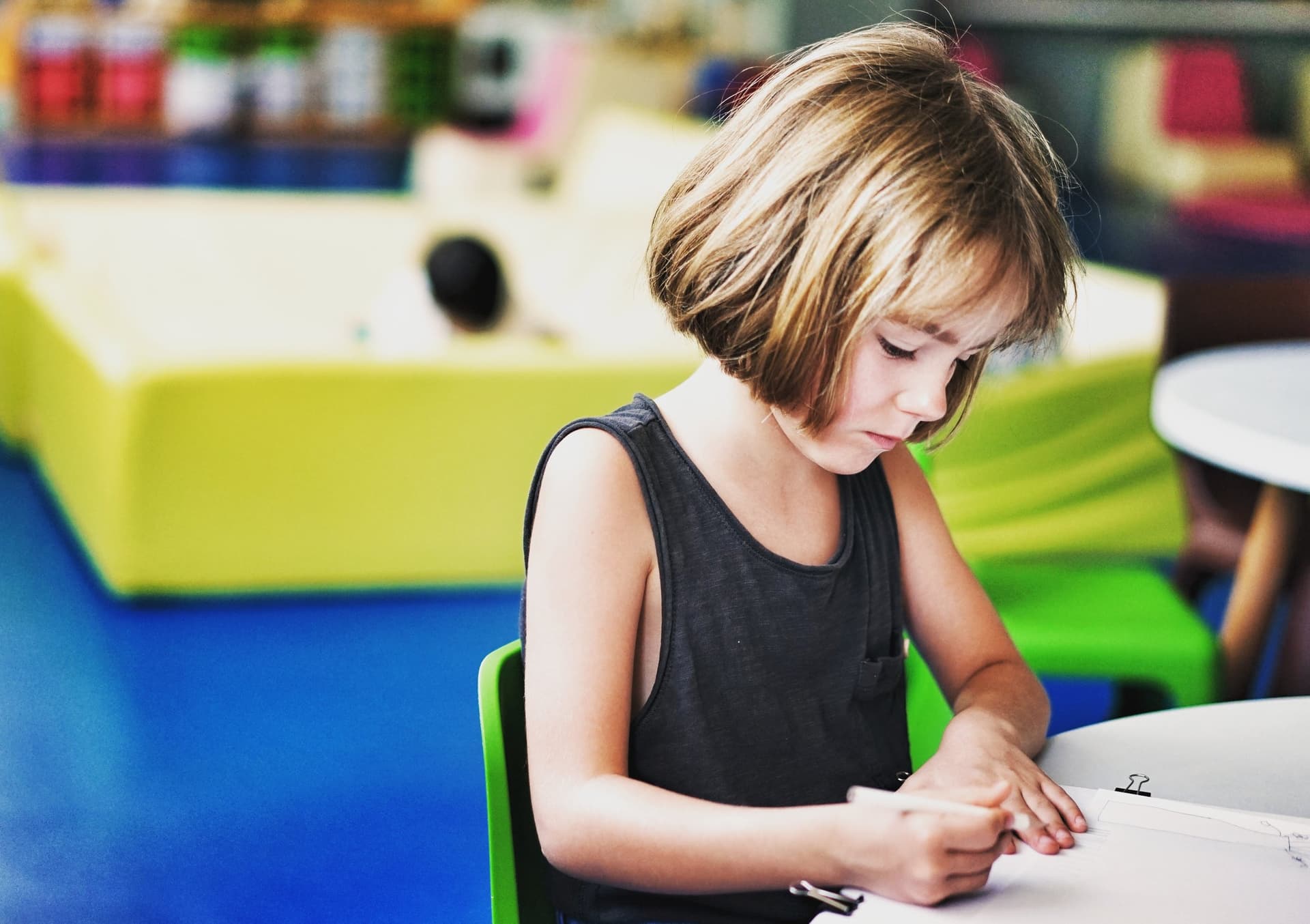 Young student in black sleeveless top concentrating on writing at desk in colorful classroom setting