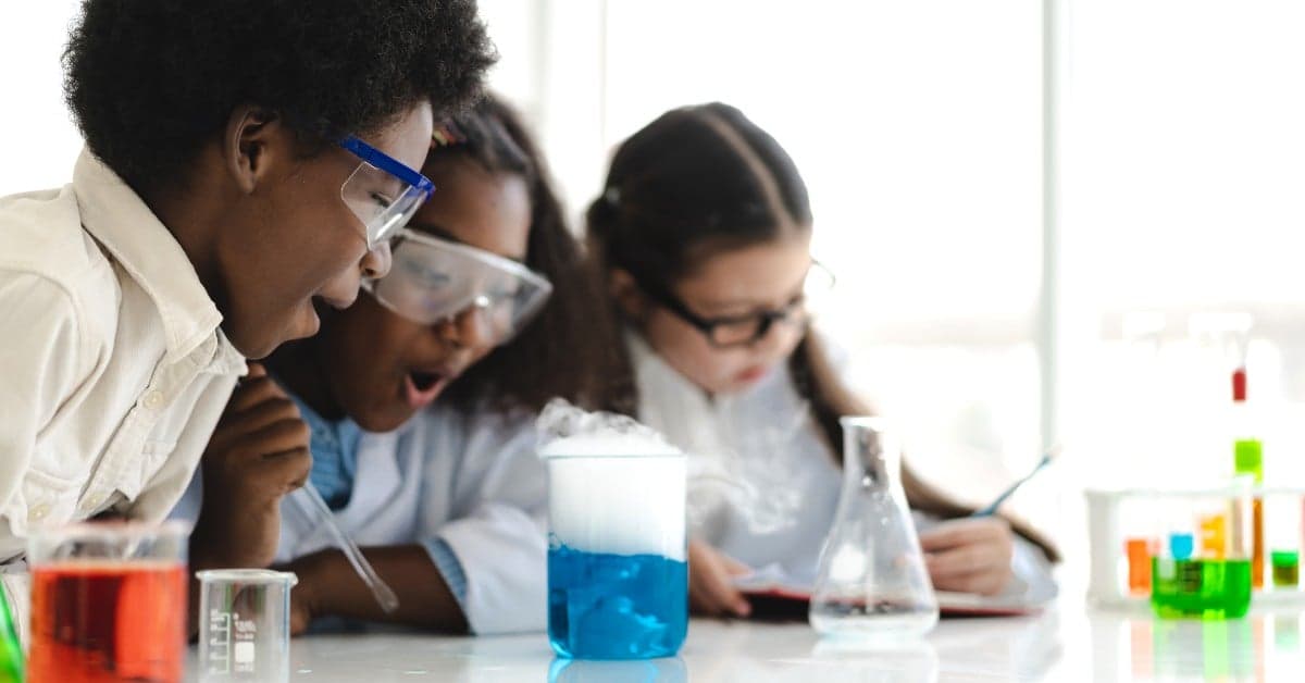 Three students in safety glasses observing a bubbling blue chemical reaction in a science laboratory