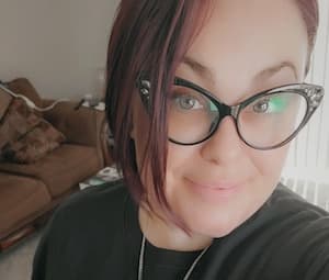 Close-up selfie of a woman with auburn hair wearing cat-eye glasses and a dark top, smiling warmly at the camera