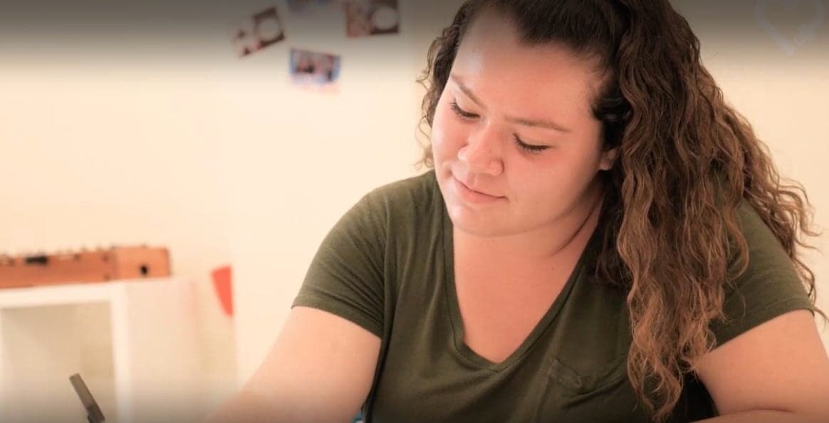 Young woman with curly hair in green t-shirt studying or writing, looking down with a gentle smile in a bright room