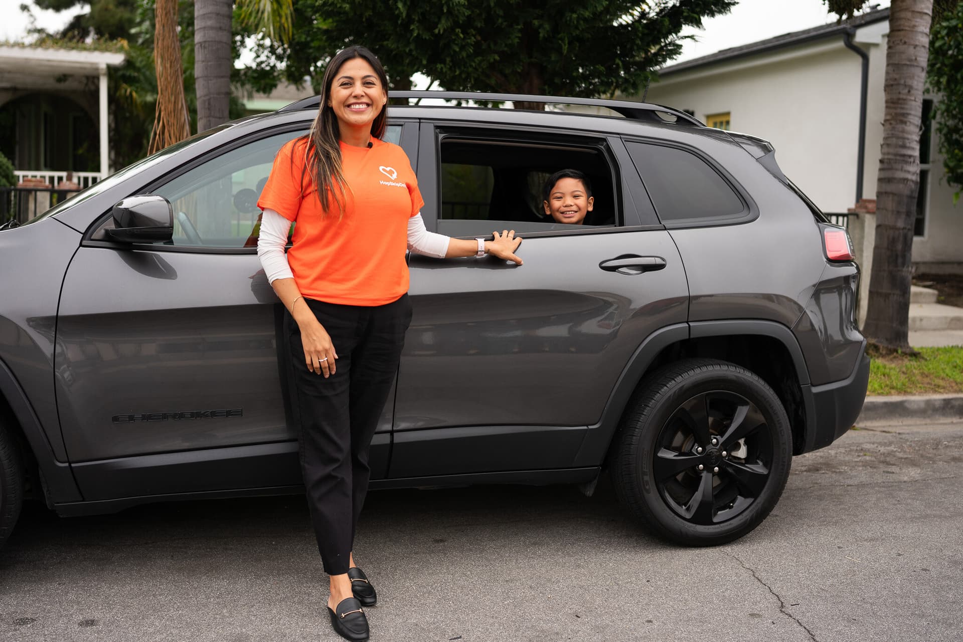 Driver in orange company shirt standing beside grey SUV with young passenger visible in back window