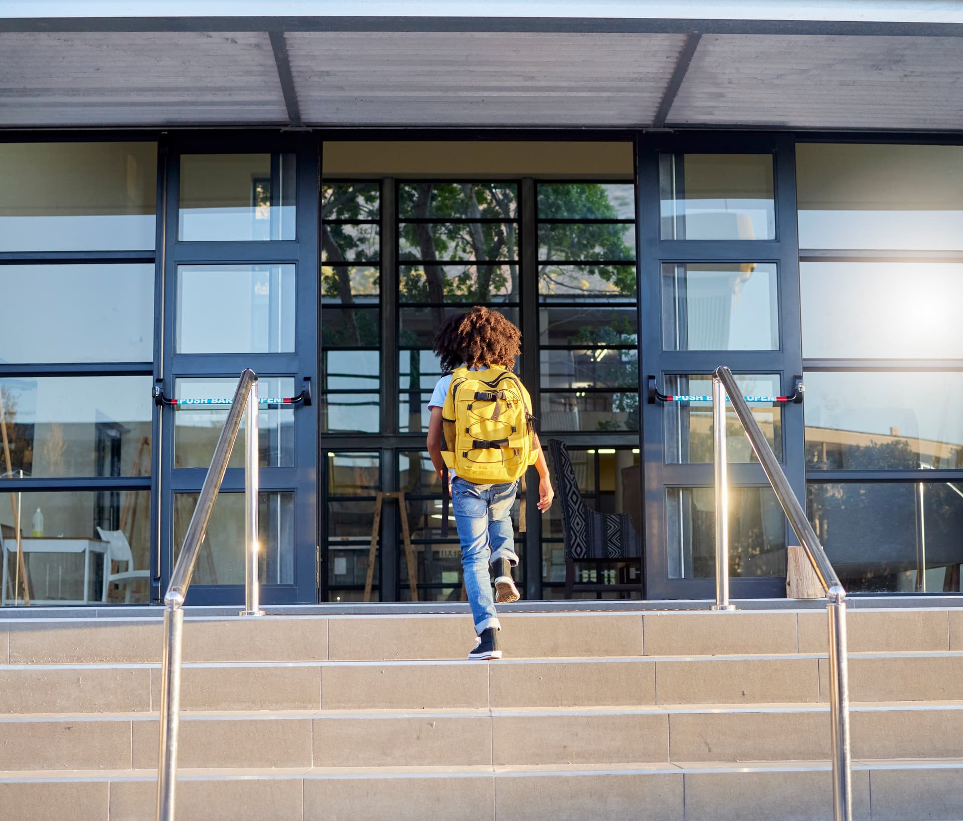 Student wearing a bright yellow backpack climbing steps toward modern glass school entrance doors