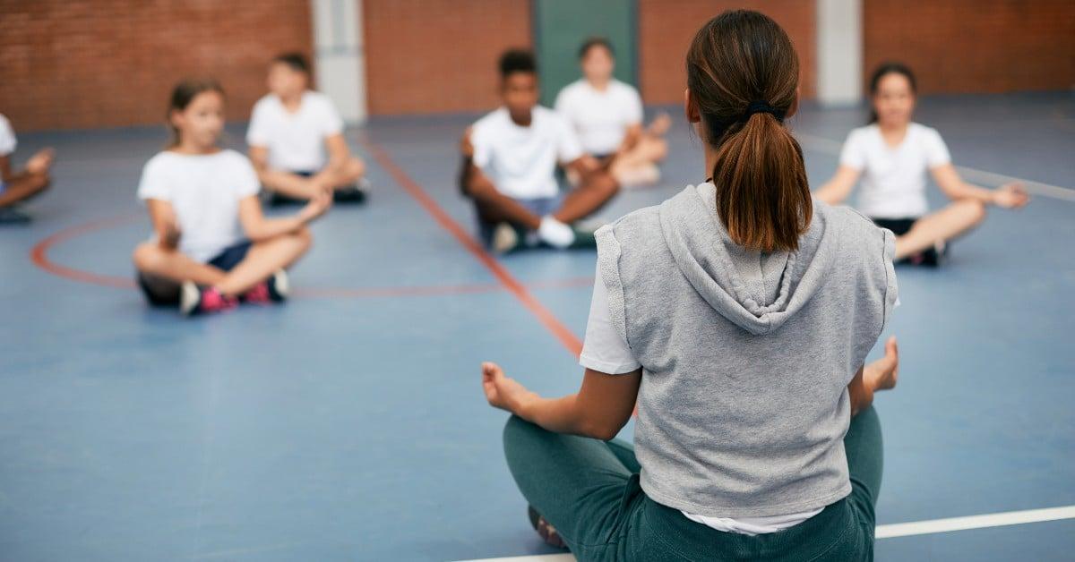 Instructor leading students in meditation session on gym floor, students sitting cross-legged in white uniforms