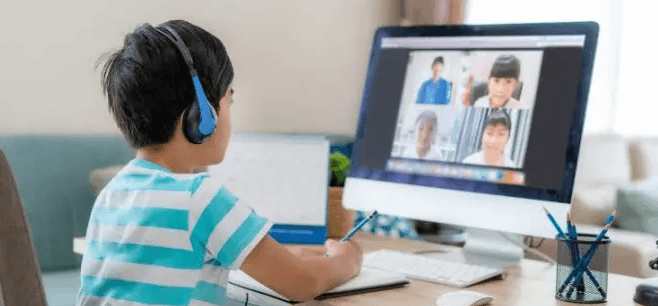 Young student wearing headphones participating in online class via video conference while taking notes at home desk