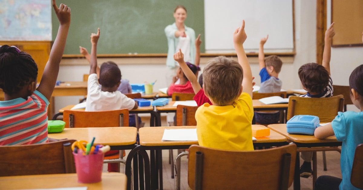 Elementary school students eagerly raising hands in classroom while teacher stands at chalkboard