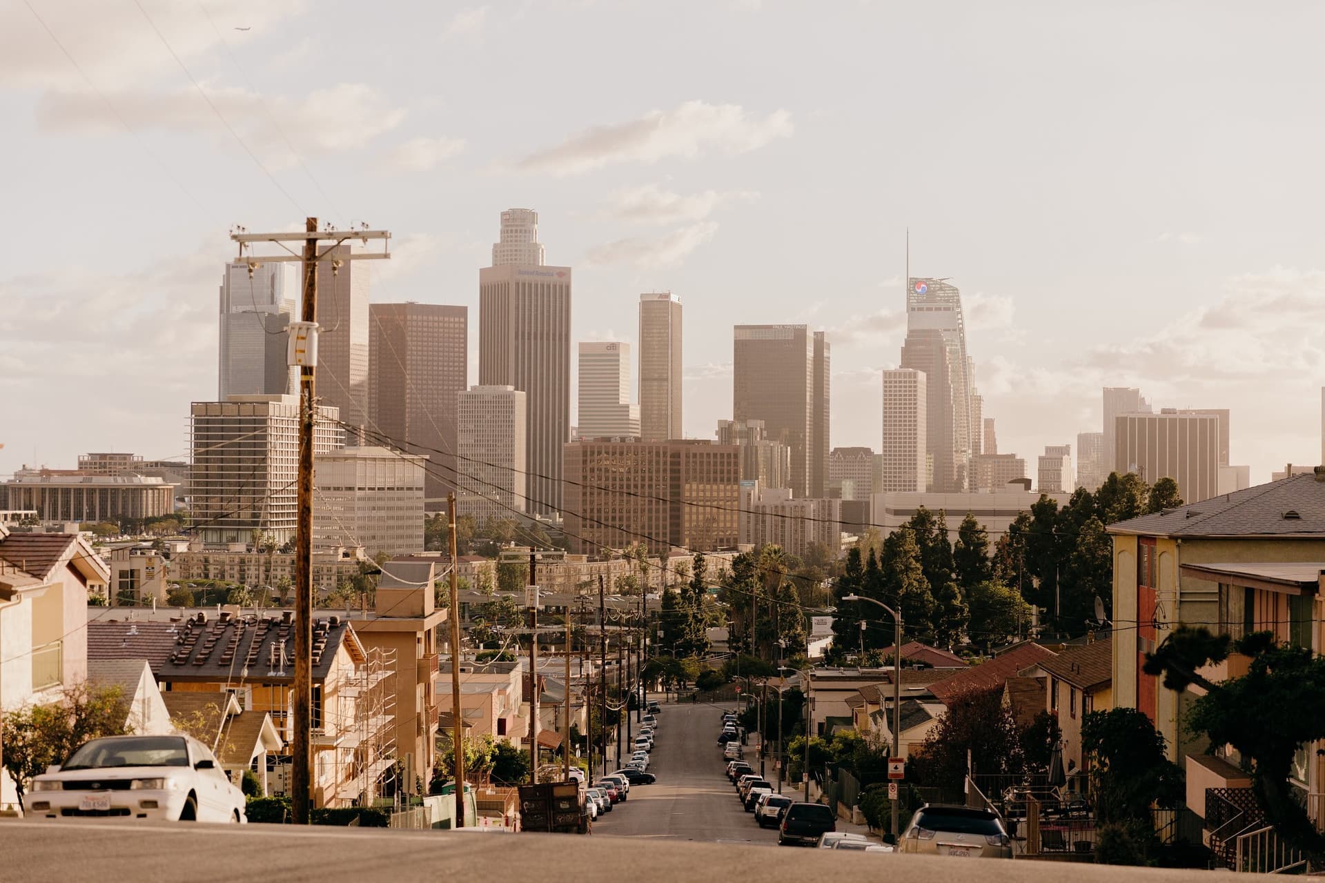 Downtown Los Angeles skyline viewed from a hillside residential street with parked cars and utility poles in the foreground