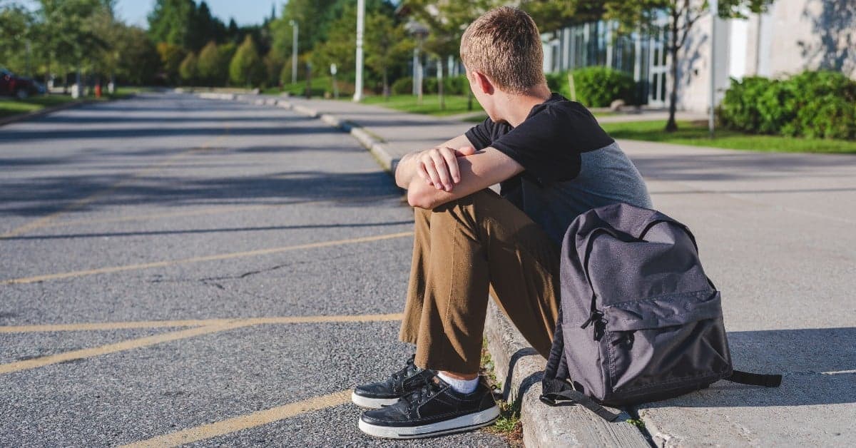 Student sitting alone on parking lot curb with backpack, viewed from behind in contemplative pose