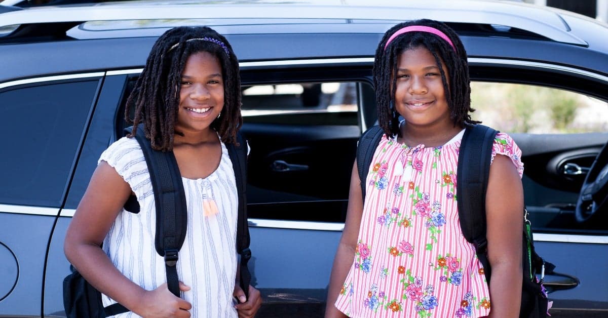 Two young students with backpacks standing by a car, wearing summer dresses and bright smiles
