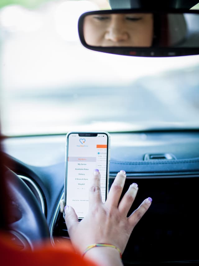 Hand reaching for smartphone mounted on car dashboard displaying a mobile app interface, with rearview mirror visible above
