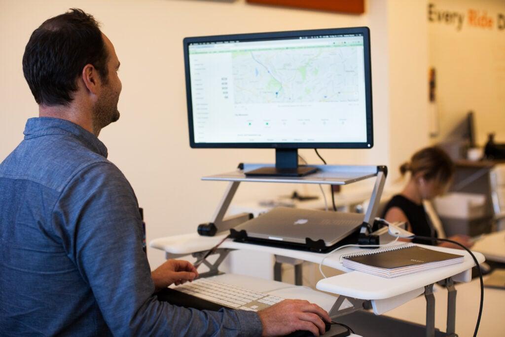 Professional working at ergonomic standing desk setup with monitor displaying map interface in modern office environment