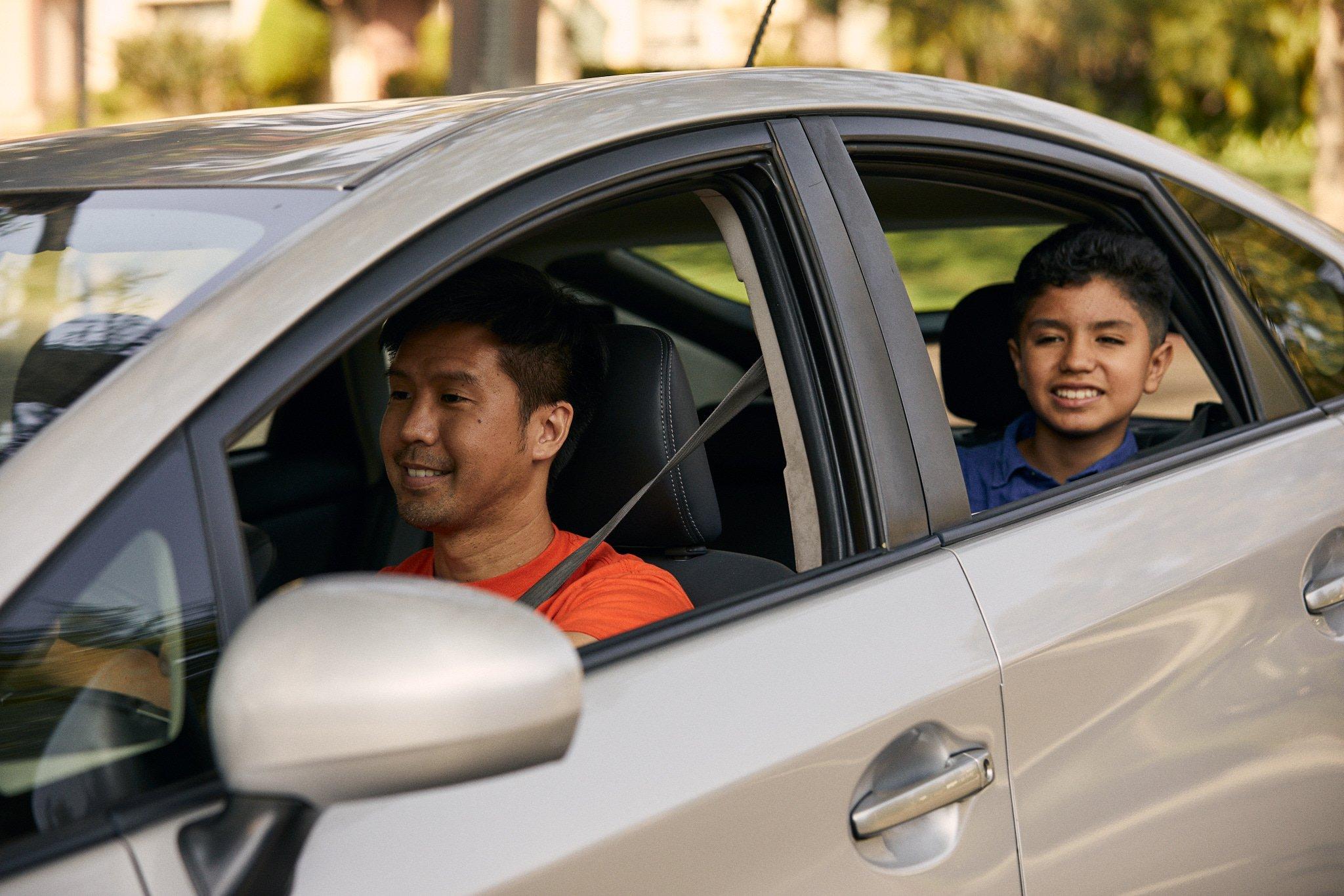 Adult driver in orange shirt and young passenger in blue shirt sitting in silver car, both smiling warmly