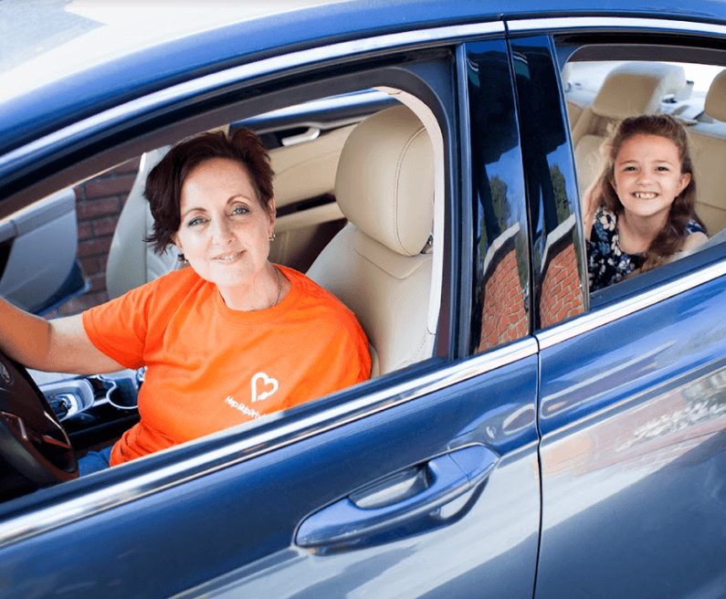 Driver in orange charity t-shirt and young passenger in back seat of blue car, both smiling at camera
