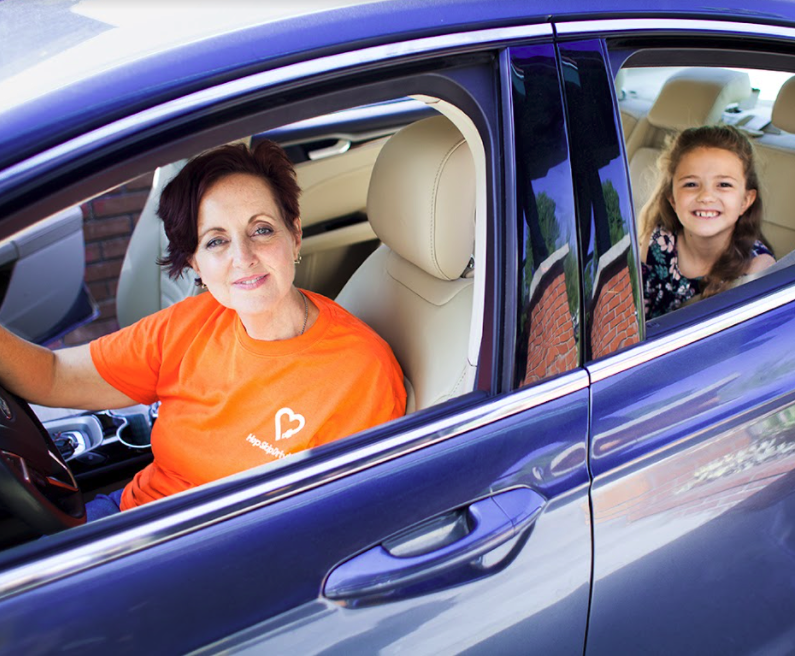 Driver in orange charity t-shirt and young passenger in back seat of blue car, both smiling at camera