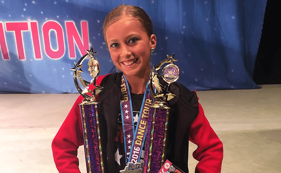 Young dancer smiling while holding two large competition trophies at a dance event with 'ITION' visible in background