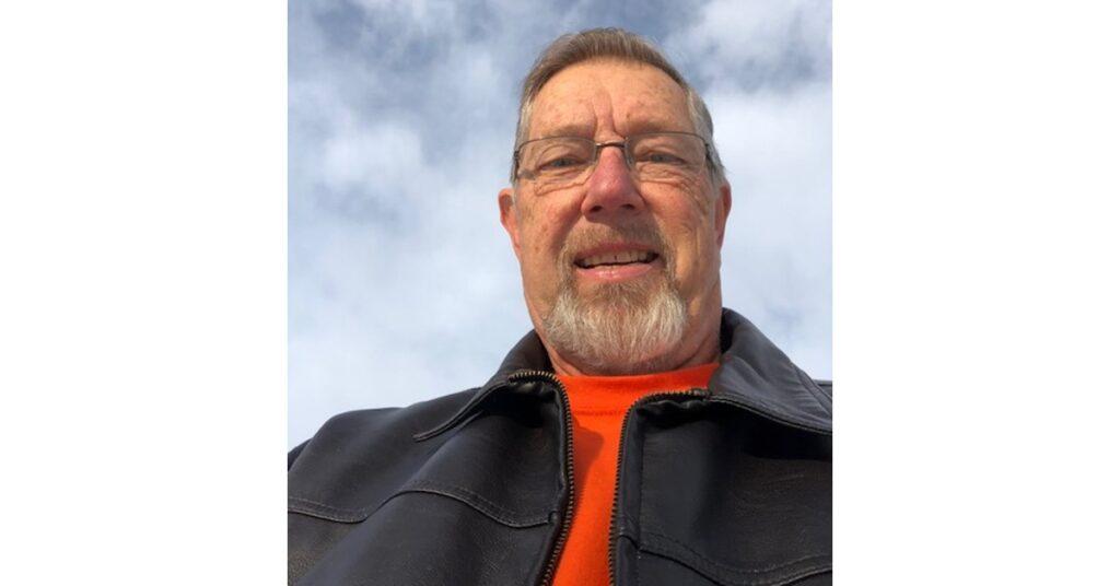 Senior man in glasses and leather jacket with bright orange shirt smiling at camera against cloudy sky background