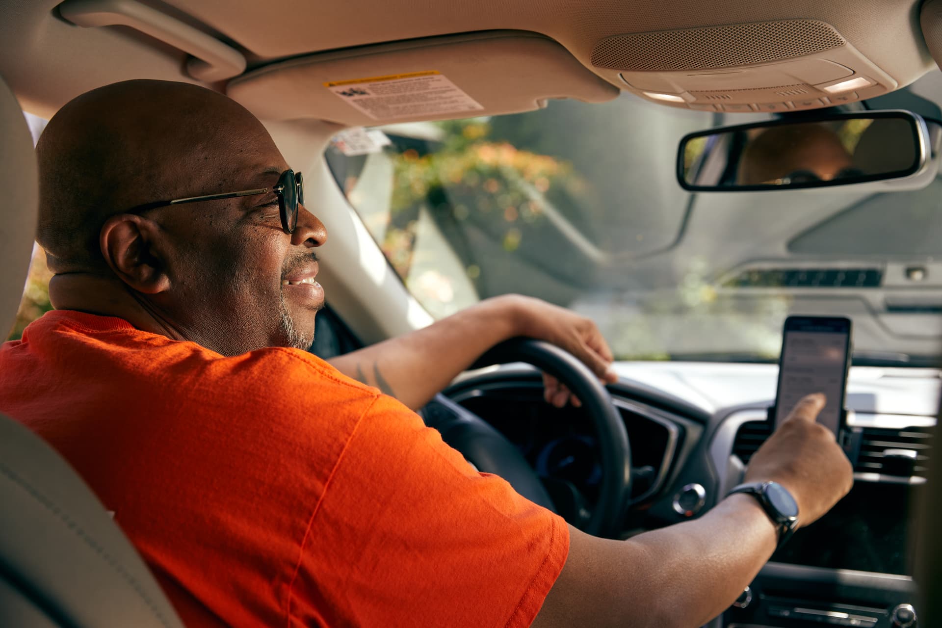 Driver in orange shirt checking smartphone while operating vehicle, seen from passenger side angle