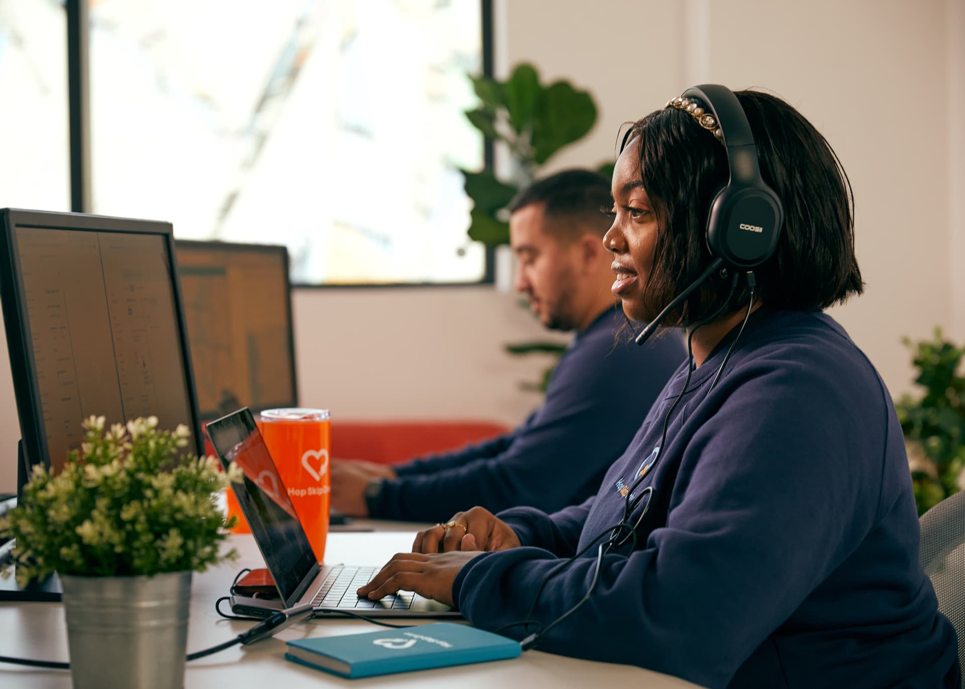 Professional wearing headset working at desk with laptop in modern office space with plants and coworkers in background