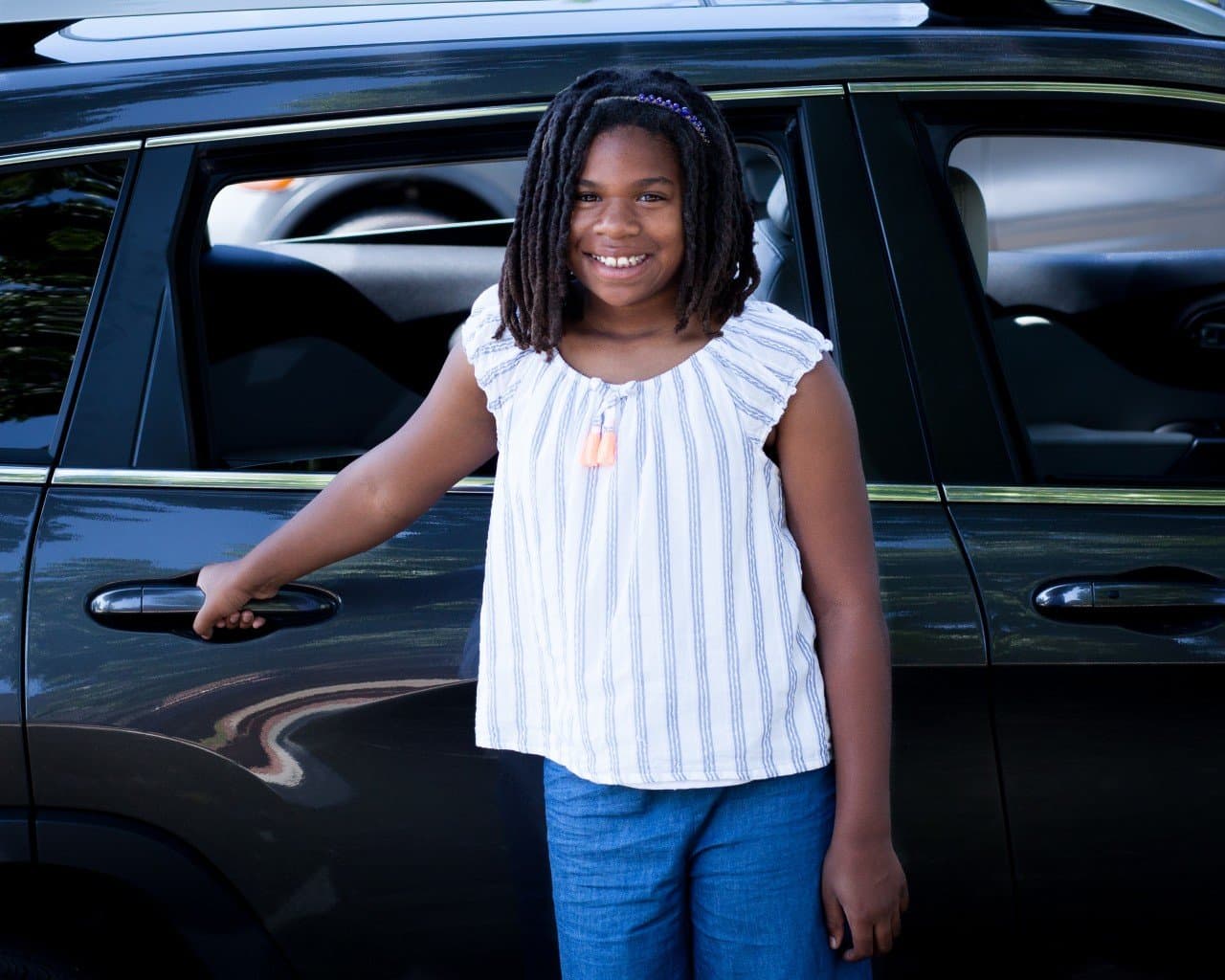 Young girl wearing striped white top and jeans standing by black car door, smiling warmly at camera