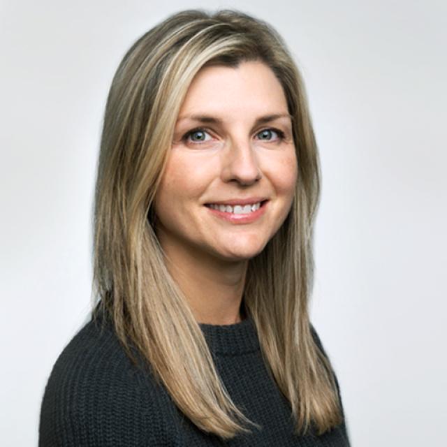 Professional headshot of a person with blonde hair wearing a black sweater, smiling warmly against a light background