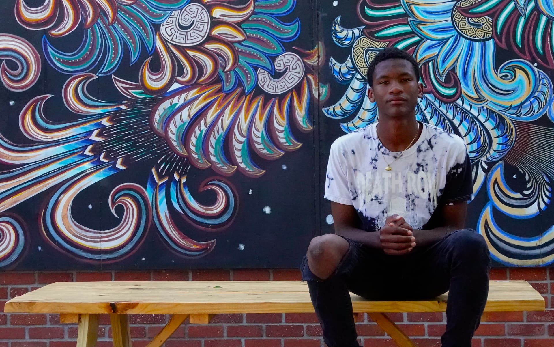 Person sitting on wooden bench against colorful swirling street art mural featuring blue and orange patterns