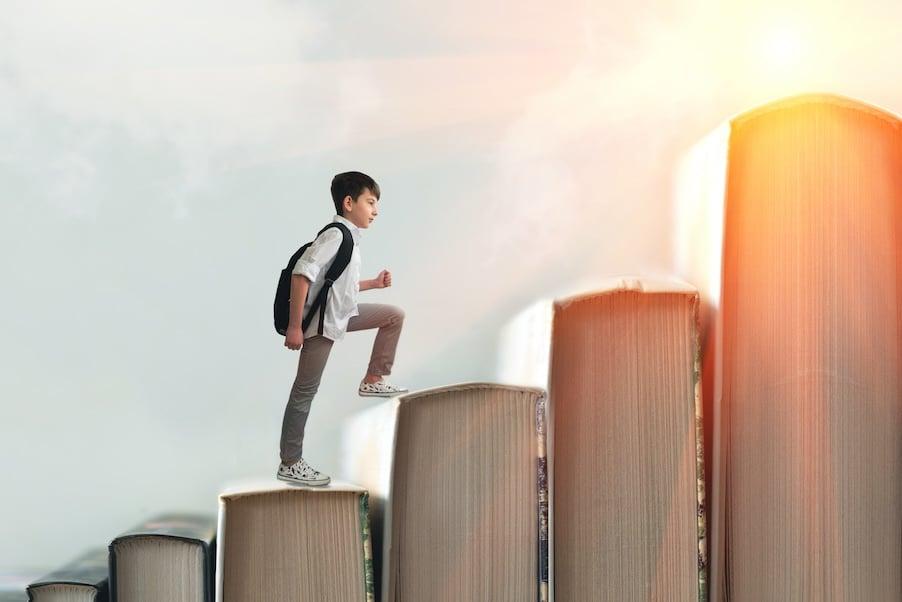Student with backpack climbing upward on giant books arranged as ascending steps against bright sky with sunlight