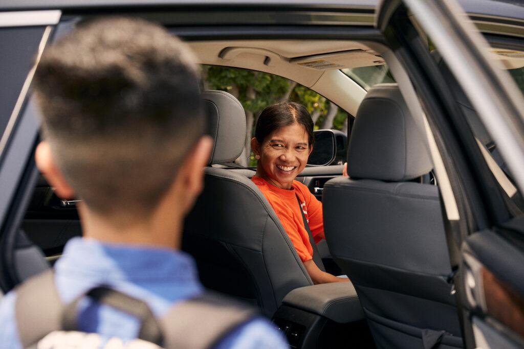 Passenger in bright orange shirt smiling while sitting in car backseat, with partial view of driver in foreground