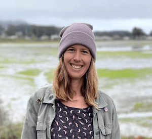 Woman wearing beanie and jacket smiling warmly against backdrop of steaming geothermal pools