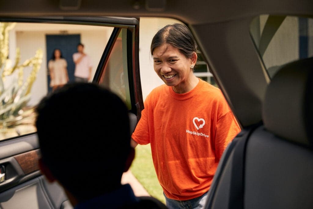 Driver in bright orange HopSkipDrive t-shirt smiling warmly while interacting with a passenger through a car door