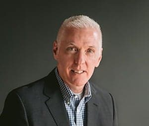 Professional headshot of person in dark suit jacket and checkered shirt against dark grey background